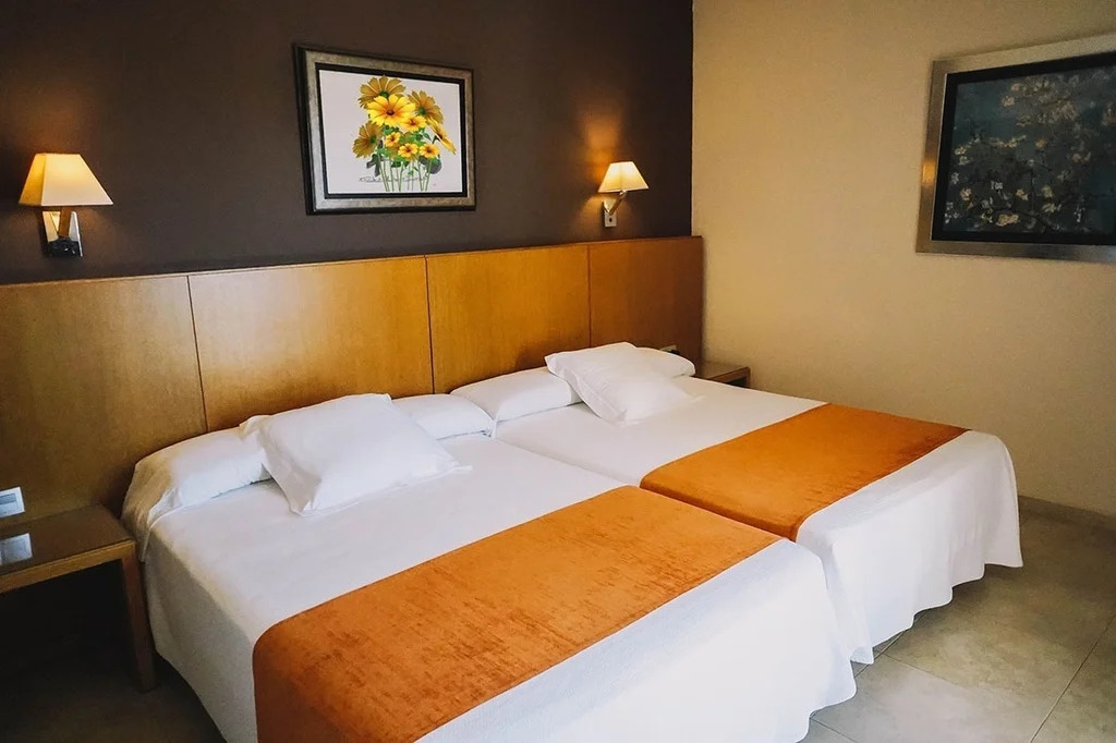 Gemütliches Hotelzimmer mit zwei Einzelbetten, dekoriert mit orangefarbenen Bettüberwürfen, einem Wandgemälde mit Sonnenblumen und warmem Licht von Wandlampen, das eine einladende Atmosphäre schafft.