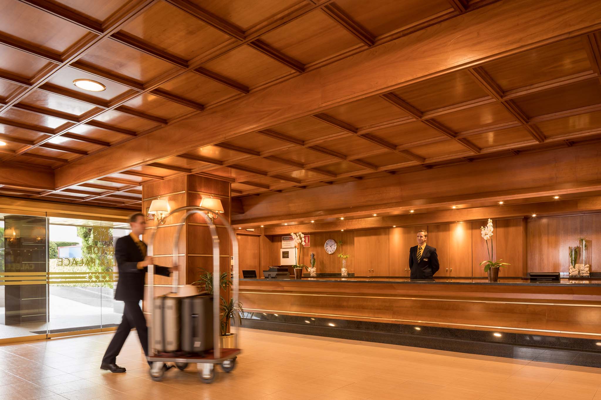 Ein einladender Blick auf die Lobby des H·TOP Calella Palace, in der warmes Holzdekor und sanfte Beleuchtung für eine gemütliche Atmosphäre sorgen. Ein Hotelangestellter in formeller Kleidung schiebt einen Gepäckwagen mit Koffern vorbei, während ein weiterer Mitarbeiter an der Rezeption steht. Im Hintergrund sind Pflanzen und dekorative Elemente zu sehen, die das elegante Ambiente unterstreichen.