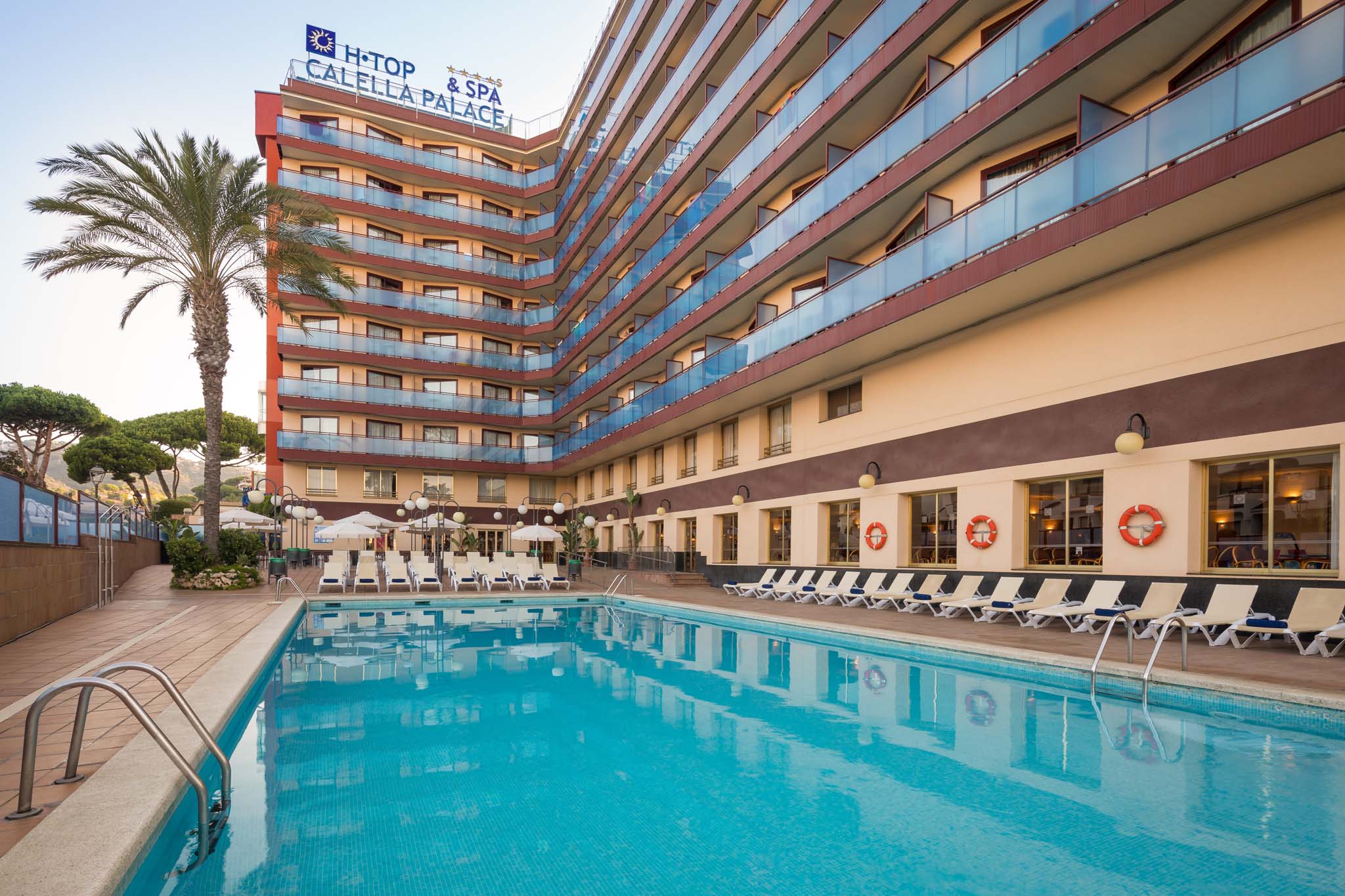 Eine eindrucksvolle Ansicht des Poolbereichs im H·TOP Calella Palace, mit einem klaren, blauen Pool, der das Licht der Abendsonne reflektiert. Die Fassade des Hotels ist farbenfroh und modern gestaltet, während Palmen und Liegen mit Sonnenschirmen den einladenden Außenbereich ergänzen. In der Nähe des Pools sind Lebensrettungsringe sichtbar, die Sicherheit und Erholung für die Gäste gewährleisten.