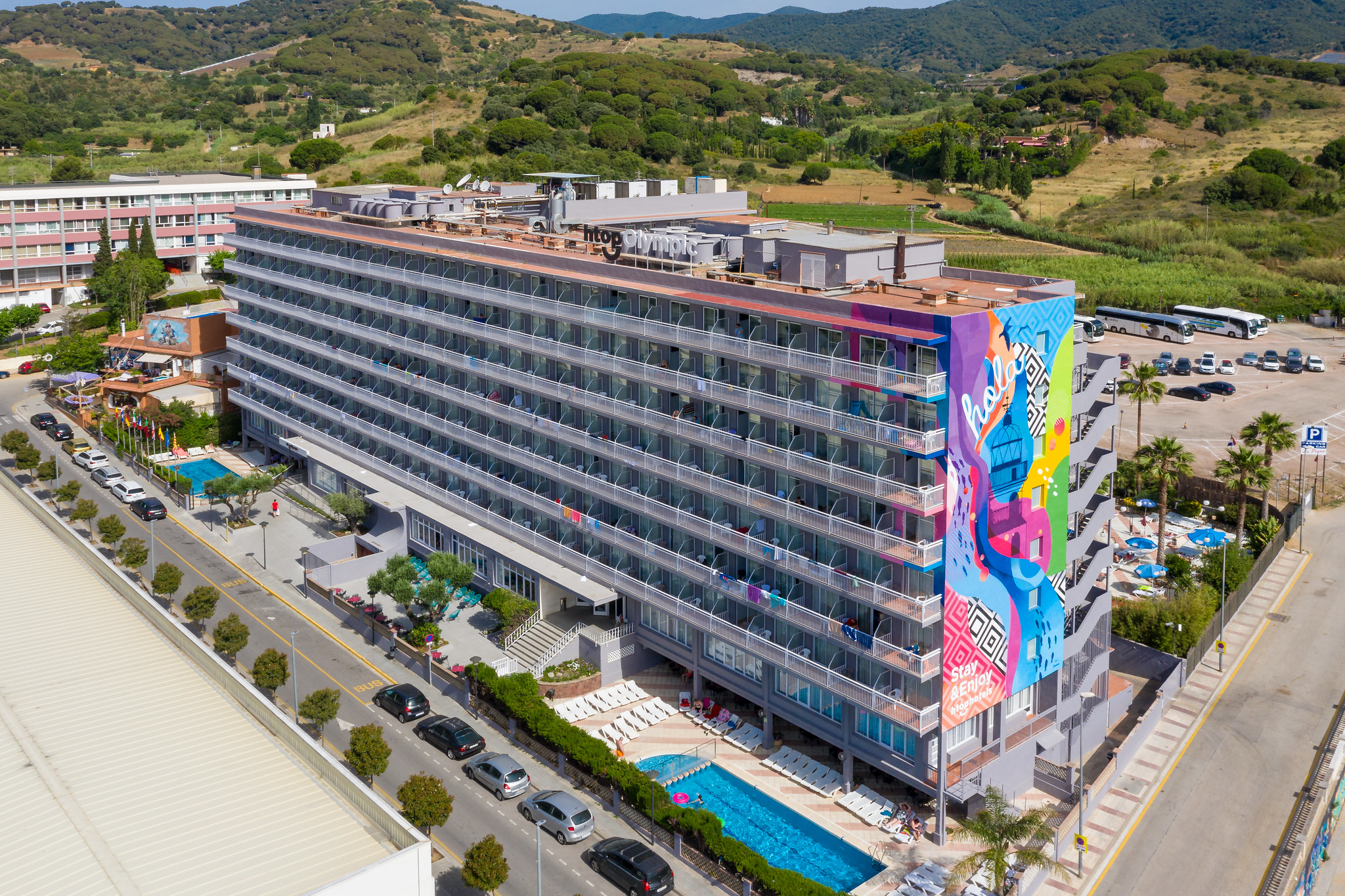 Eine Gesamtansicht des htops Olympic Hotels, die die stilvolle Architektur und die große, bunte Wandkunst zeigt, die die Fassade schmückt. Im Vordergrund sind die gepflegten Gärten und der Swimmingpool des Hotels zu sehen, die eine einladende Umgebung schaffen.