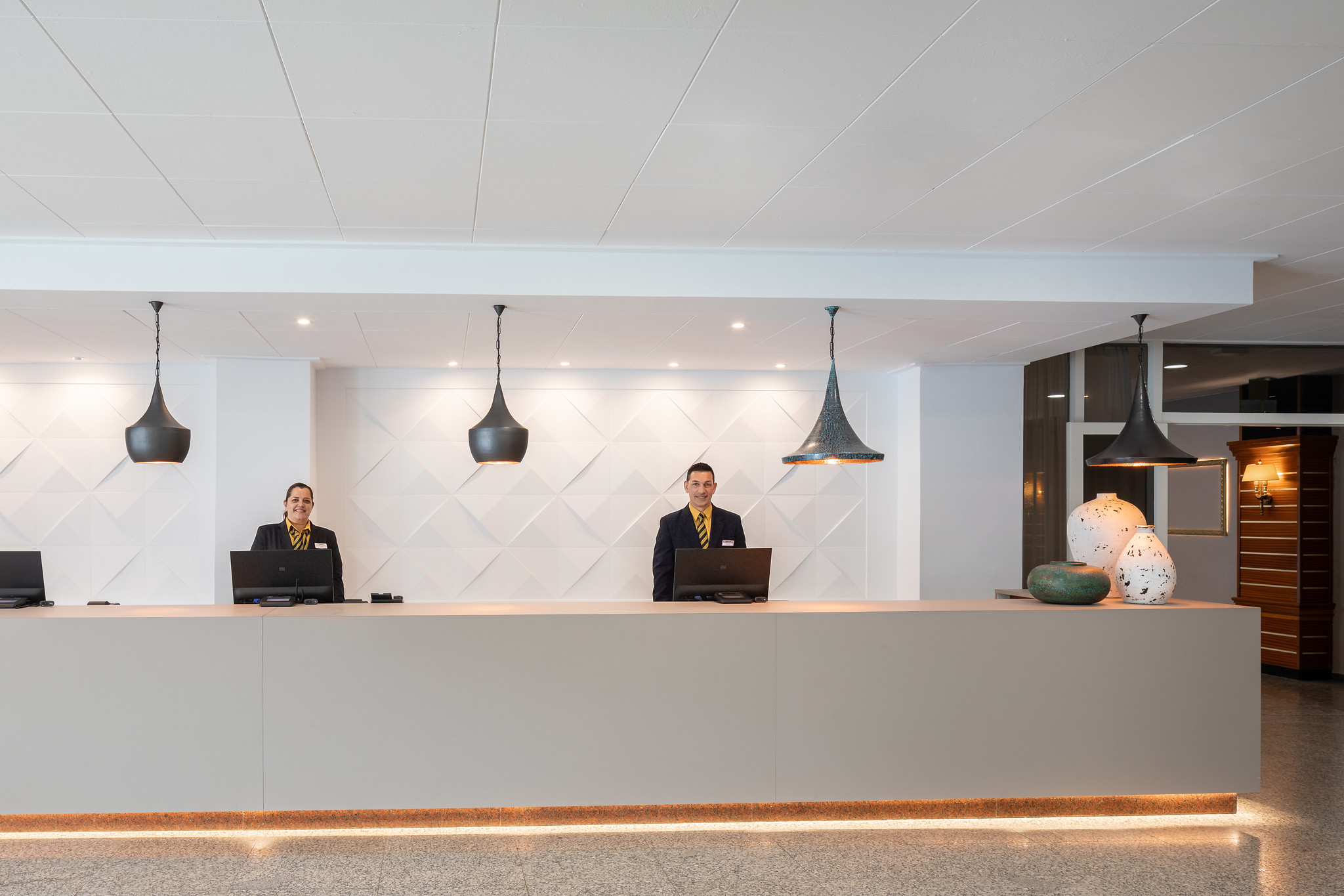 Der Empfangsbereich des Hotel Olympic, modern und einladend gestaltet, wird von zwei freundlichen Empfangsmitarbeitern betreut. Die Empfangstheke aus hellem Material und der elegante Hintergrund mit geometrischem Muster schaffen eine stilvolle Atmosphäre. Über der Theke hängen zwei moderne, schwarze Pendelleuchten, die sanftes Licht spenden. Die Mitarbeiter sind in formeller Kleidung und wirken bereit, den Gästen bei ihren Anfragen zu helfen. Auf der Theke stehen Computerbildschirme, und im Hintergrund sind dekorative Elemente, darunter stilvolle Vasen, sichtbar, die dem Raum einen Hauch von Eleganz verleihen.