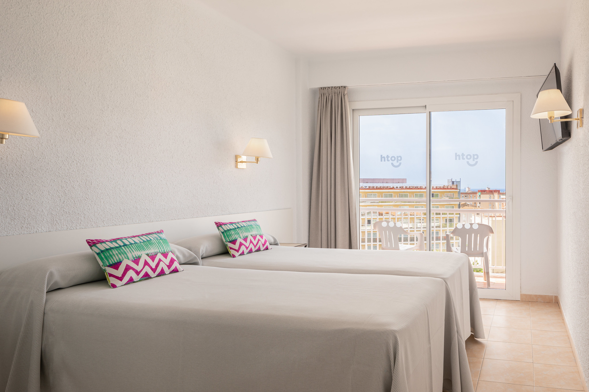 Eine helle und einladende Hotelzimmeransicht, die zwei Einzelbetten mit modernen, bunten Kissen zeigt. Die Wände sind in einem sanften Weiß gehalten, während das natürliche Licht durch die große Balkontür strömt, die einen Blick auf den Balkon und die umliegenden Gebäude bietet. Elegante Wandlampen fügen eine warme Atmosphäre hinzu, und ein Fernseher ist an der Wand montiert, um den Komfort der Gäste zu erhöhen.
