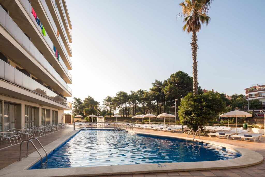 Der Poolbereich des Htop Royal Star & SPA in Lloret de Mar, umgeben von Sonnenschirmen und Liegen, bietet eine entspannende Atmosphäre. Die moderne Architektur des Hotels und die grüne Umgebung laden zu einem erholsamen Tag am Wasser ein.