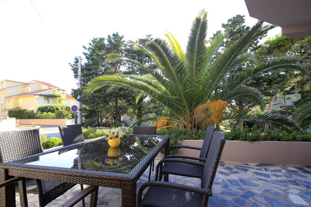 Gemütliche Terrasse mit einem eleganten Tisch und Stühlen, umgeben von grünen Pflanzen und Palmen, ideal für entspannende Momente im Freien in den Komfort Apartments.